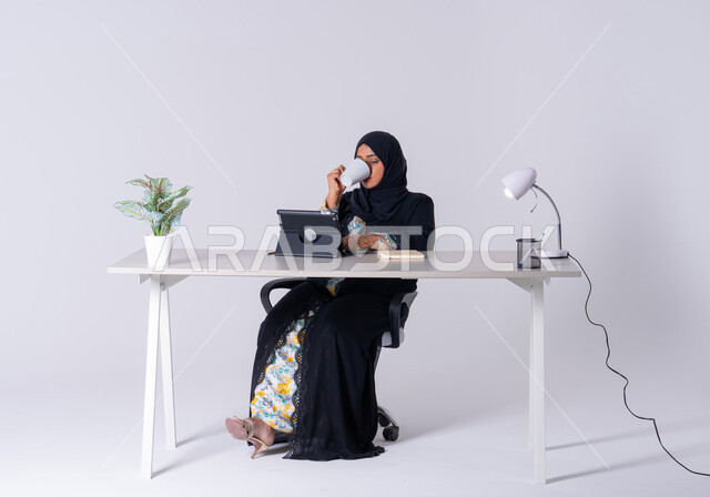 تدوين وتسجيل الملاحظات والمعلومات على الورق, سيده اماراتيه تقوم بإدارة الأعمال عن بعد وتنظيمها،  بورتريه لامراه عربيه خليجيه سعوديه ترتدي العباءه والحجاب تجلس على مكتب ،تعمل على جهاز التابلت ،استخدم التقنيات الحديثة في الأعمال الإدارية ،كتابة مهام واهداف العمل ، احتساء المشروبات الساخنه