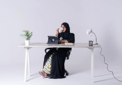 Taking notes and recording information on paper, an Emirati woman managing and organizing business remotely, a portrait of a Saudi Arabian Gulf woman wearing an abaya and hijab sitting at a desk, working on a tablet, using modern technologies in administrative work, writing down work tasks and goals, drinking hot drinks