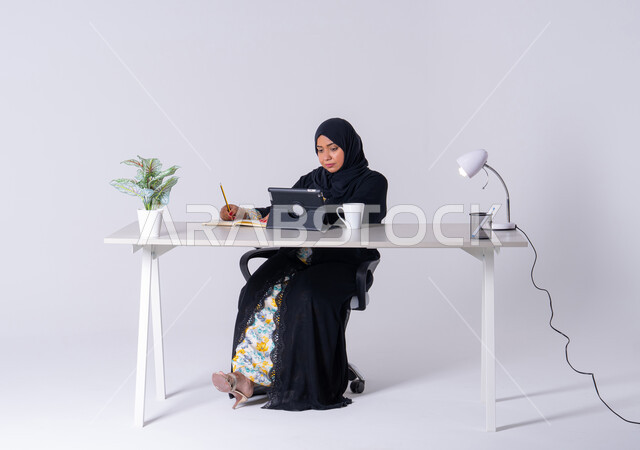 Emirati woman managing and organizing business remotely, taking notes and recording information on paper, portrait of a Saudi Arabian Gulf woman wearing an abaya and hijab sitting at a desk, working on a tablet, using modern technologies in administrative work, writing down work tasks and goals, drinking hot drinks