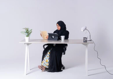 Use modern technologies in administrative work, an Emirati woman manages and organizes business remotely, taking notes and recording information on paper, a portrait of a Saudi Arabian Gulf woman wearing an abaya and hijab sitting at a desk, working on a tablet, writing down work tasks and goals, and drinking hot drinks