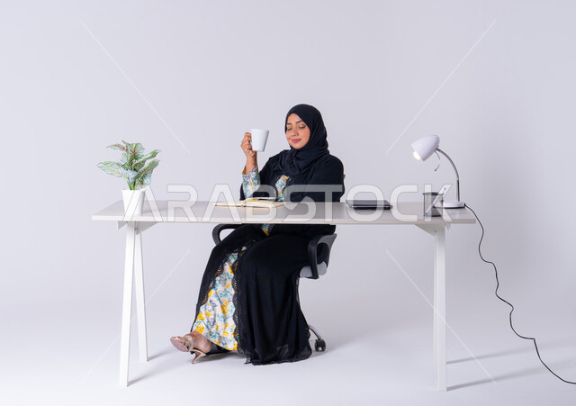تدوين وتسجيل الملاحظات والمعلومات على الورق, استخدام الاوراق البيضاء الفارغة في العمل، احتساء المشروبات الساخنه، سيده اماراتيه تقوم بإدارة الأعمال وتنظيمها، بورتريه لامراه عربيه خليجيه سعوديه ترتدي العباءه والحجاب تجلس على مكتب ، وتقوم بكتابة المهام والأهداف اليومية