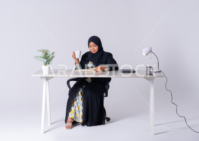استخدام الاوراق البيضاء الفارغة في العمل، احتساء المشروبات الساخنه، سيده اماراتيه تقوم بإدارة الأعمال وتنظيمها،  تدوين وتسجيل الملاحظات والمعلومات على الورق, بورتريه لامراه عربيه خليجيه سعوديه ترتدي العباءه والحجاب تجلس على مكتب ، وتقوم بكتابة المهام والأهداف اليومية
