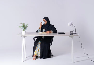 Drinking hot drinks, Emirati woman managing and organizing business, using blank white papers at work, writing down and recording notes and information on paper, portrait of a Saudi Arabian Gulf woman wearing an abaya and hijab sitting at a desk, writing down daily tasks and goals