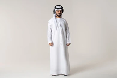 Attention to appearance, entertainment by listening to audio clips with headphones, following music and entertainment with headphones, wearing audio headsets, a portrait of an Emirati Gulf Arab man wearing the Emirati kandura listening to songs with gestures of joy and happiness, white background.