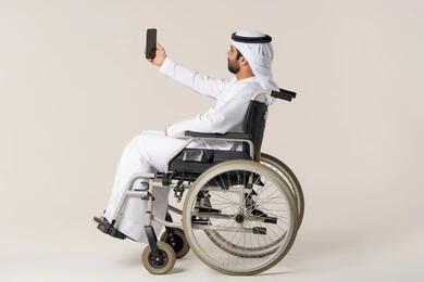 Preserving happy moments through photos, the concept of elegance, masculinity, and attention to appearance, a side portrait of an Emirati Arab man with special needs wearing a kandura and a white ghutrah sitting in a wheelchair holding a mobile phone and taking a selfie, gestures of self-confidence, white background.