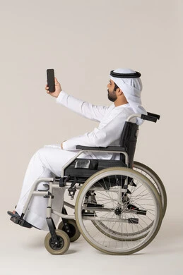 Capturing distinctive shots to document memories, the concept of elegance, masculinity, and attention to appearance, a side portrait of an Emirati Arab man with special needs wearing a kandura and a white ghutrah sitting in a wheelchair holding a mobile phone and taking a selfie, gestures of self-confidence, white background.