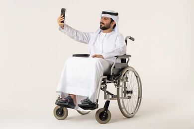 The concept of elegance, masculinity, and attention to appearance, documenting happy moments and taking commemorative photos, a portrait of an Arab Gulf Emirati man with special needs wearing a kandura and a white ghutrah sitting in a wheelchair holding a mobile phone and taking a selfie, gestures of self-confidence, white background.