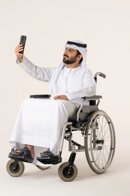 توثيق اللحظات السعيدة والتقاط الصور التذكارية, بورترية لرجل عربي خليجي اماراتي من ذوى الاحتياجات الخاصة يرتدي الدشداشة و الغترة البيضاء يجلس على كرسي متحرك يمسك الجوال ويقوم بأخذ صورة سيلفي لنفسه, إيماءات الثقة بالنفس, مفهوم الأناقة والرجولة والاهتمام بالمظهر الخارجي, خلفية بيضاء