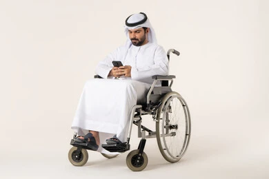 تجربة التسوق الإلكتروني باستخدام تطبيقات الجوال، بورترية لرجل عربي خليجي اماراتي من ذوى الاحتياجات الخاصة يرتدي الدشداشة و الغترة البيضاء يجلس على كرسي متحرك يحمل الهاتف المحمول، تطور التكنولوجيا والأجهزة التقنية، الدردشة مع الأهل والأصدقاء، إيماءات الاندماج والتركيز، خلفية بيضاء