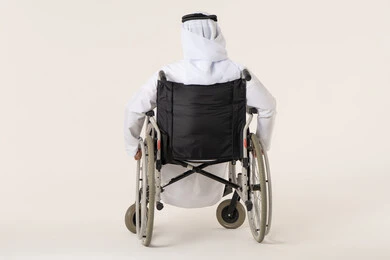 تحسين جودة حياة ذوي الاعاقه الحركيه، استخدام كراسي المعاقين للمساعده على الحركه والتنقل, بورترية من الخلف لرجل عربي خليجي اماراتي من ذوى الاحتياجات الخاصة يرتدي الدشداشة و الغترة البيضاء يجلس على كرسي متحرك، خلفيه بيضاء