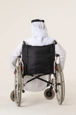 استخدام كراسي المعاقين للمساعده على الحركه والتنقل, تحسين جودة حياة ذوي الاعاقه الحركيه, بورترية من الخلف لرجل عربي خليجي اماراتي من ذوى الاحتياجات الخاصة يرتدي الدشداشة و الغترة البيضاء يجلس على كرسي متحرك، خلفيه بيضاء