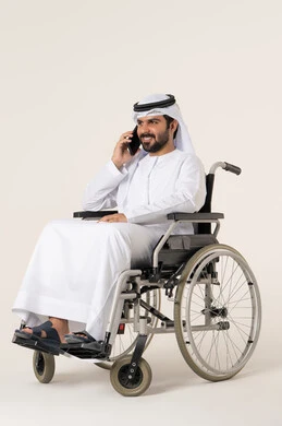 Enjoying exchanging conversations and news, making a voice phone call via mobile, a portrait of a young Emirati Arab man with special needs wearing a kandura and a white ghutrah sitting in a wheelchair and talking on a mobile phone, gestures of happiness in communicating with friends and relatives, white background.