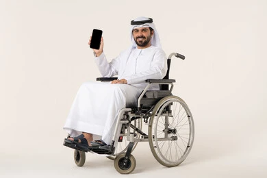 الاستخدام اليومي للتقنيات الحديثة, النظر إلى الكاميرا بإيماءات السعادة والسرور, بورترية لشاب عربي خليجي اماراتي من ذوى الاحتياجات الخاصة يرتدي الدشداشة و الغترة البيضاء يجلس على كرسي متحرك و يمسك بيده الهاتف المحمول, عرض شاشة سوداء فارغة عبر الجوال, خلفية بيضاء