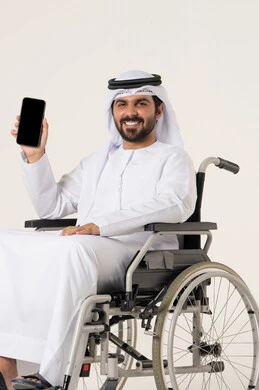 استخدام الأجهزة التقنية الحديثة في الحياة اليومية, النظر إلى الكاميرا بإيماءات السعادة والسرور, بورترية لشاب عربي خليجي اماراتي من ذوى الاحتياجات الخاصة يرتدي الدشداشة و الغترة البيضاء يجلس على كرسي متحرك و يمسك بيده الهاتف المحمول, عرض شاشة سوداء فارغة عبر الجوال, خلفية بيضاء