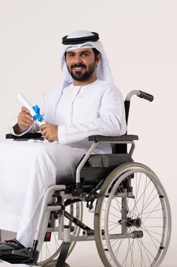 Celebrating the completion of university studies, the concept of success and excellence, looking at the camera with gestures of joy and satisfaction, a portrait of a young Arab Gulf Emirati man with special needs wearing a kandura and a white ghutrah sitting in a wheelchair holding his graduation certificate, with a white background.