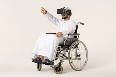 الاشارة الى شيء ما، الاندماج ومحاكاة العالم الخيالي، المتعة والتسلية بالمشاهدة عبر نظارة الواقع الافتراضي، بورترية لشاب عربي خليجي اماراتي من ذوى الاحتياجات الخاصة يرتدي الزي التقليدي و نظارات ثلاثية الأبعاد (الفي ار) يجلس على كرسي متحرك, التطور التقني والتكنولوجي ، خلفية بيضاء
