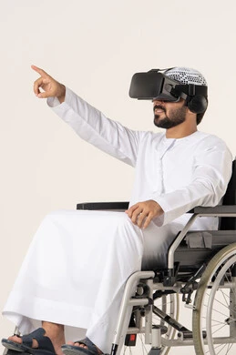The enjoyment and entertainment of watching through virtual reality glasses, a portrait of a young Arab Gulf Emirati man with special needs wearing traditional attire and 3D glasses (VR) sitting in a wheelchair, the integration and simulation of the imaginary world, technological and technical development, pointing to something, white background.