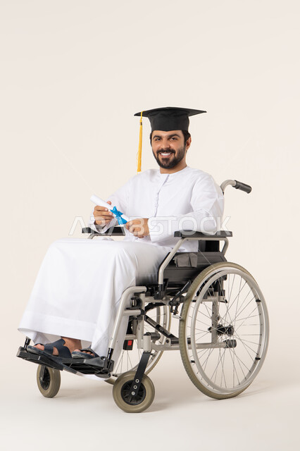 الاحتفاء بالنجاح الأكاديمي والتفوق الدراسي، مفهوم الاجتهاد والتفوق، بورتريه لطالب عربي اماراتي خليجي من ذوي الاحتياجات الخاصة يرتدي الثوب الأبيض وقبعة التخرج يجلس على كرسي متحرك يمسك بيده الشهادة , النظر الى الكاميرا بإيماءات الرضا والسرور