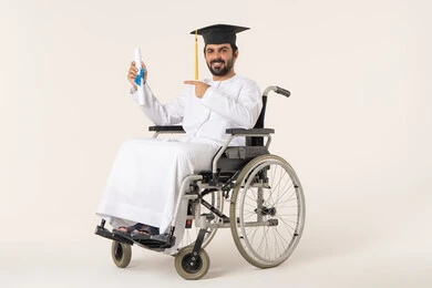 Celebrating the completion of university studies, the concept of diligence and excellence, a portrait of an Emirati Arab Gulf student with special needs wearing a white gown and graduation cap sitting in a wheelchair holding a certificate and pointing to it, looking at the camera with gestures of satisfaction and joy.