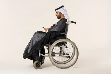 التحدث والدردشة مع الأصدقاء عبر الجوال، ايماءات الاندماج والتركيز، تصفح وسائل التواصل الاجتماعي، بورتريه من الجانب لرجل عربي خليجي اماراتي من ذوي الاحتياجات الخاصة يرتدي الثوب التقليدي يجلس على كرسي متحرك يستخدم الهاتف المحمول، خلفية بيضاء