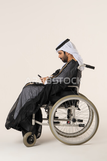 ايماءات الاندماج والتركيز، تصفح وسائل التواصل الاجتماعي، بورتريه من الجانب لرجل عربي خليجي اماراتي من ذوي الاحتياجات الخاصة يرتدي الثوب التقليدي يجلس على كرسي متحرك يستخدم الهاتف المحمول, التحدث والدردشة مع الأصدقاء عبر الجوال، خلفية بيضاء