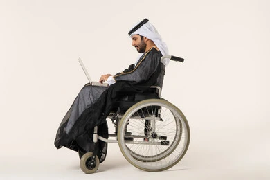 متابعة سير العمل عن بعد , استخدام الأجهزة والتقنيات الحديثة, الاقتصاد والأمور المالية، بورتريه من الجانب لرجل عربي خليجي اماراتي من ذوي الاحتياجات الخاصة يرتدي الثوب التقليدي يجلس على كرسي متحرك يعمل على الحاسوب المحمول، خلفية بيضاء