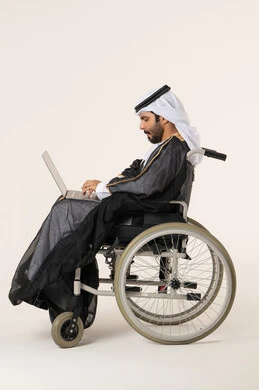 إنجاز المهام الإدارية عبر الإنترنت , استخدام الأجهزة والتقنيات الحديثة, الاقتصاد والأمور المالية، بورتريه من الجانب لرجل عربي خليجي اماراتي من ذوي الاحتياجات الخاصة يرتدي الثوب التقليدي يجلس على كرسي متحرك يعمل على الحاسوب المحمول، خلفية بيضاء