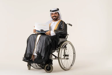 لحظة التخرج من الجامعة، مفهوم الاجتهاد والتفوق، الشعور بالفرح والفخر والثقة بالنفس، بورتريه لطالب عربي اماراتي خليجي من ذوي الاحتياجات الخاصة يرتدي الثوب التقليدي يجلس على كرسي متحرك يمسك بيده شهادة التخرج, النظر الى الكاميرا بإيماءات الرضا والسرور, خلفية بيضاء
