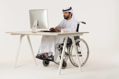 متابعة تحقيق الخطط المستقبلية، إيماءات الاندماج والتركيز أثناء العمل، مهن ووظائف ادارية مكتبية, إدارة شؤون الأعمال وتنظيمها، بورتريه لرجل عربي خليجي اماراتي من ذوي الاحتياجات الخاصة يرتدي الثوب التقليدي يجلس على كرسي متحرك يعمل على الحاسوب المحمول، خلفية بيضاء