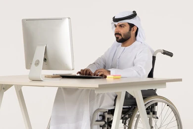 Gestures of engagement and focus while working, working to achieve goals and strategies, administrative and office professions and jobs, managing and organizing business affairs, a portrait of an Emirati Arab man with special needs wearing traditional attire sitting in a wheelchair working on a laptop, white background.