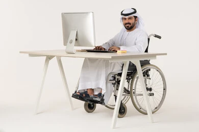 Looking at the camera with gestures of happiness and joy, working to achieve goals and strategies, administrative and office professions, managing and organizing business affairs, a portrait of an Emirati Arab man with special needs wearing traditional attire sitting in a wheelchair working on a laptop, white background.
