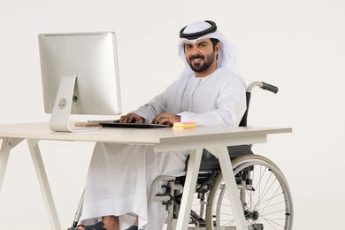 العمل لتحقيق الاهداف والاستراتيجيات، مهن ووظائف ادارية مكتبية, إدارة شؤون الأعمال وتنظيمها، بورتريه لرجل عربي خليجي اماراتي من ذوي الاحتياجات الخاصة يرتدي الثوب التقليدي يجلس على كرسي متحرك يعمل على الحاسوب المحمول, النظر الى الكاميرا بايماءات السعادة و السرور، خلفية بيضاء