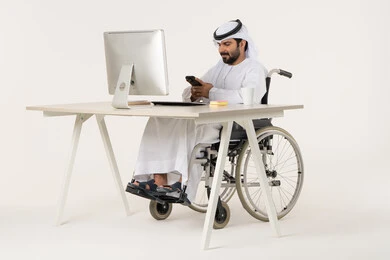 دمج التكنولوجيا المتطورة بالحياة اليومية، تصفح مواقع التواصل الاجتماعي، الدردشة مع الأهل والأصدقاء، استخدام جهاز تقني حديث، بورتريه لرجل عربي خليجي اماراتي من ذوي الاحتياجات الخاصة يرتدي الثوب التقليدي يجلس على كرسي متحرك امام الحاسوب المحمول, خلفية بيضاء