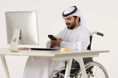 Browsing social media, chatting with family and friends, using modern technology, integrating advanced technology into daily life, a portrait of an Arab Gulf Emirati man with special needs wearing traditional attire sitting in a wheelchair in front of a laptop, white background.