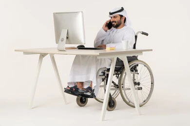 Making a phone call via mobile, communicating with family and friends, using modern technologies, a portrait of an Emirati Arab man with special needs wearing traditional attire sitting in a wheelchair in front of a laptop, gestures of happiness and joy, white background.