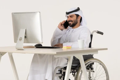 Communication with family and friends, making a phone call via mobile, using modern technologies, a portrait of an Emirati Arab man with special needs wearing traditional attire sitting in a wheelchair in front of a laptop, gestures of happiness and joy, white background.