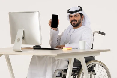 A display of a blank black screen, the use of modern technologies, the quality of mobile applications and services, a portrait of an Emirati Arab man with special needs wearing traditional attire sitting in a wheelchair in front of a laptop, looking at the camera with gestures of happiness and joy, white background.