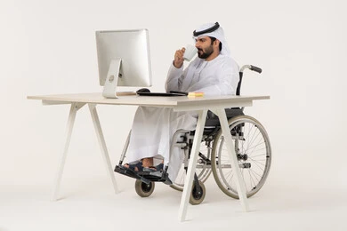 إدماج الوسائل التقنية في المهام الوظيفية، العمل على الحاسوب المحمول داخل المكتب، مهن ووظائف ادارية مكتبية، إدارة شؤون الأعمال وتنظيمها عبر الاجهزة التقنية، الاستمتاع بتناول مشروبات، بورتريه لرجل عربي خليجي اماراتي من ذوي الاحتياجات الخاصة يرتدي الثوب التقليدي يجلس على كرسي متحرك , خلفية بيضاء