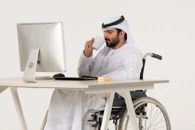 العمل على الحاسوب المحمول داخل المكتب، دمج العمل مع التقنية والتكنولوجيا، مهن ووظائف ادارية مكتبية، إدارة شؤون الأعمال وتنظيمها عبر الاجهزة التقنية، بورتريه لرجل عربي خليجي اماراتي من ذوي الاحتياجات الخاصة يرتدي الثوب التقليدي يجلس على كرسي متحرك , الاستمتاع بتناول المشروبات , خلفية بيضاء