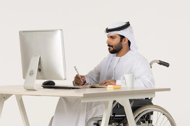 Writing and recording information on paper, writing daily tasks and goals, managing and organizing business affairs through technical devices, a portrait of an Arab Gulf Emirati man with special needs wearing traditional attire sitting in a wheelchair in front of a laptop, white background.