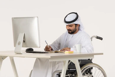 كتابة المهام والأهداف اليومية, تدوين وتسجيل المعلومات على الورق، إدارة شؤون الأعمال وتنظيمها عبر الاجهزة التقنية، بورتريه لرجل عربي خليجي اماراتي من ذوي الاحتياجات الخاصة يرتدي الثوب التقليدي يجلس على كرسي متحرك امام الحاسب المحمول , خلفية بيضاء