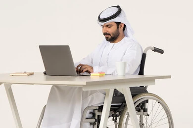 دمج العمل مع التقنية والتكنولوجيا، مهن ووظائف ادارية مكتبية، إدارة شؤون الأعمال وتنظيمها عبر الاجهزة التقنية، بورتريه لرجل عربي خليجي اماراتي من ذوي الاحتياجات الخاصة يرتدي الثوب التقليدي يجلس على كرسي متحرك , العمل على الحاسوب المحمول داخل المكتب, الاستمتاع بتناول المشروبات , خلفية بيضاء