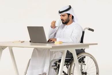 إدارة شؤون الأعمال وتنظيمها عبر الاجهزة التقنية، مهن ووظائف ادارية مكتبية، دمج العمل مع التقنية والتكنولوجيا، بورتريه لرجل عربي خليجي اماراتي من ذوي الاحتياجات الخاصة يرتدي الثوب التقليدي يجلس على كرسي متحرك , العمل على الحاسوب المحمول داخل المكتب, الاستمتاع بتناول المشروبات , خلفية بيضاء