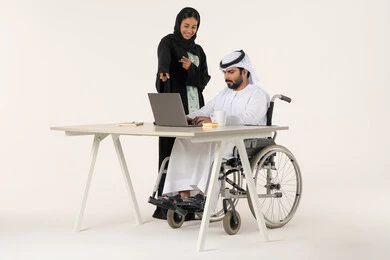 تحقيق اهداف الشركه, بورتريه لرجل عربي خليجي اماراتي من ذوي الاحتياجات الخاصة يرتدي الثوب التقليدي يجلس على كرسي متحرك يستخدم الحاسب المحمول , امراة خليجيه مبتسمه تقوم بالاشارة الى شيء ما , خلفية بيضاء