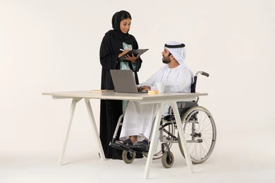 الوحدة و العمل الجماعي, بورتريه لرجل عربي خليجي اماراتي من ذوي الاحتياجات الخاصة يرتدي الثوب التقليدي يجلس على كرسي متحرك يستخدم الحاسب المحمول , توزيع مهام العمل, امراة خليجيه مبتسمه تمسك بيديها حامل الاوراق , خلفية بيضاء