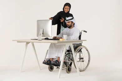 Achieving the company's goals, the Emirati work environment, unity and teamwork, a portrait of an Arab Gulf Emirati man with special needs wearing traditional attire sitting in a wheelchair using a laptop, a smiling Gulf woman pointing at something, white background.