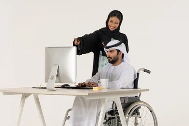 بيئة العمل الاماراتي , الوحده والعمل الجماعي, تحقيق اهداف الشركه, بورتريه لرجل عربي خليجي اماراتي من ذوي الاحتياجات الخاصة يرتدي الثوب التقليدي يجلس على كرسي متحرك يستخدم الحاسب المحمول , امراة خليجيه مبتسمه تقوم بالاشارة الى شيء ما , خلفية بيضاء