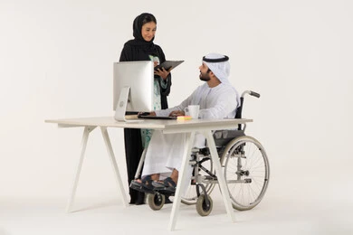 مناقشة عمل بين زملاء عمل ,   بورتريه لرجل عربي خليجي اماراتي من ذوي الاحتياجات الخاصة يرتدي الثوب التقليدي يجلس على كرسي متحرك يستخدم الحاسب المحمول , توزيع مهام العمل, امراة خليجيه مبتسمه تمسك بيديها حامل الاوراق , خلفية بيضاء