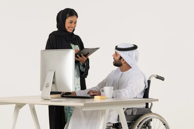 بيئة العمل الاماراتي, بورتريه لرجل عربي خليجي اماراتي من ذوي الاحتياجات الخاصة يرتدي الثوب التقليدي يجلس على كرسي متحرك يستخدم الحاسب المحمول , توزيع مهام العمل, امراة خليجيه مبتسمه تمسك بيديها حامل الاوراق , خلفية بيضاء