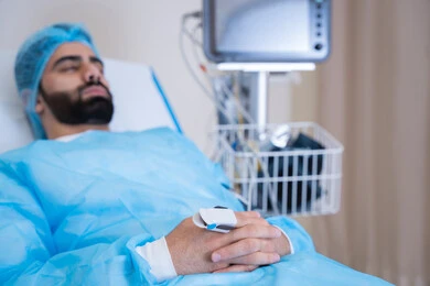 An intensive care room showing an Emirati Gulf Arab patient lying on a hospital bed under medical supervision. The patient's pulse is monitored using an oximeter. Medical tools and equipment are also available.
